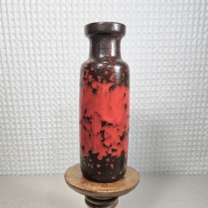 May include: A cylindrical ceramic vase featuring a dark brown base color with a prominent, textured bright red glaze pattern covering the center. The vase has a narrow neck and a flared rim, standing upright on a circular wooden base.