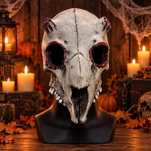 Peut inclure: Un masque de crâne d'animal beige avec deux petites cornes sur le dessus, des orbites profondes soulignées de rouge foncé et une rangée de dents blanches le long de la mâchoire. Le masque est présenté sur un support noir, avec une texture osseuse.
