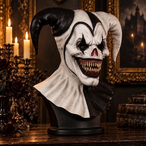 Puede incluir: Un busto de máscara de bufón en blanco y negro con una amplia sonrisa dentada, cuernos puntiagudos y marcas faciales dramáticas. El tocado está dividido en colores contrastantes, con una cara blanca texturizada y una base oscura y sólida.