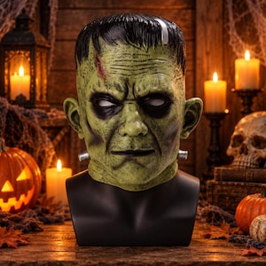 Peut inclure: Un masque de monstre de Frankenstein vert avec une coiffure noire à sommet plat, une cicatrice frontale proéminente, des yeux enfoncés et des boulons argentés dépassant du cou, présenté sur un buste de mannequin noir lisse.