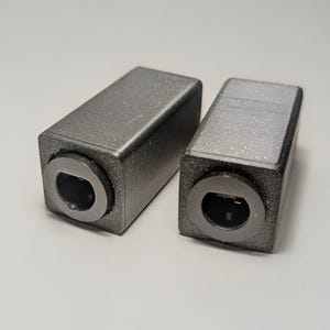 Pode incluir: Duas carcaças retangulares de plástico cinza-escuro impressas em 3D com acabamento de superfície texturizado. Cada unidade apresenta uma porta circular embutida na face frontal, revelando componentes eletrônicos internos e um pequeno pino.