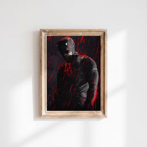 Op de afbeelding: Een ingelijste kunstdruk met een donkere, monochrome afbeelding van een gemaskerde superheld in een gestructureerd pak met een rood dubbel-D-embleem op de borst, tegen een achtergrond van verticale rode strepen die op regen lijken.
