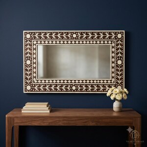 May include: A rectangular wall mirror featuring a dark brown frame with intricate white floral and geometric inlay patterns. The frame displays a repeating border of diamonds and stylized leaf motifs, surrounding a clear, reflective glass surface.