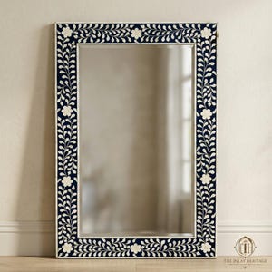 May include: A rectangular wall mirror featuring a dark navy blue frame with an intricate white floral and vine inlay pattern. The design consists of repeating leaf motifs and five-petaled flowers that border the entire reflective glass surface.