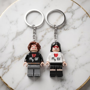 Może przedstawiać: Dwa breloki z plastikowymi minifigurkami. Lewa figurka ma brązowe włosy, czarną koszulkę z czerwonym sercem i napisem My GF oraz szare spodnie. Prawa figurka ma czarne włosy, okulary w kształcie serc, białą koszulkę z czerwonym sercem i napisem My BF oraz czarną spódnicę.