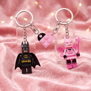 Peut inclure: Deux porte-clés avec des figurines Lego reliées par un cœur séparable. L'un comporte un Batman noir avec une cape. L'autre présente un Batman fée rose avec des ailes et une baguette étoilée. Les deux ont des anneaux et chaînes en métal argenté.