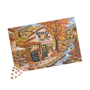 May include: A jigsaw puzzle depicting a fall scene with a Cafe & Books shop, a stone bridge over a flowing stream, and vibrant orange trees. A menu board lists Coffee, Tea, Pastries & Cakes, Soup of the Day, and Books & Reading. Loose pieces are below.