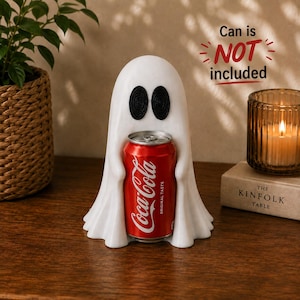 Puede incluir: Una figura blanca con forma de fantasma y dos ojos ovalados negros, diseñada para sostener una lata de bebida. La figura presenta una parte superior lisa y redondeada con bordes drapeados. El texto indica: Can is NOT included.