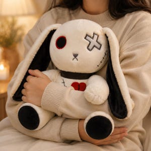 May include: A plush rabbit toy with cream-coloured fur and long ears featuring black inner linings. The face has one red circular eye, one grey cross-shaped eye, and a small nose. A red heart is stitched on the chest, and black patches cover the feet.