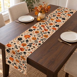 Puede incluir: Un camino de mesa rectangular largo con fondo color crema y un patrón repetitivo de calabazas naranjas, calabazas de Halloween negras y follaje oscuro. La tela muestra varias formas de calabazas, algunas con caras talladas y enredaderas.
