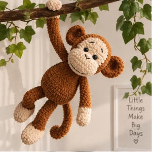 May include: A brown and cream-colored crocheted monkey plush toy with a textured yarn finish, round ears, and black eyes. The monkey is hanging from a branch by one hand, featuring light-colored hands and feet, and a long, curled tail.