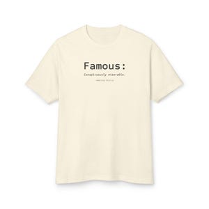 May include: A cream-colored short-sleeve t-shirt featuring black text printed on the chest. The text reads: Famous: Conspicuously miserable. -Ambrose Bierce. The shirt has a crew neckline and a standard fit, presented against a plain white background.