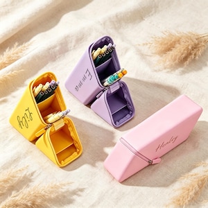 May include: Three triangular pencil cases in yellow, lavender, and light pink. The yellow case is labeled Lily, the lavender case is labeled Emma, and the pink case is labeled Henley. Each case features a zippered opening and internal compartments.