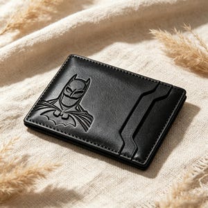 May include: A black rectangular wallet featuring an embossed Batman logo on the left side. The wallet has a smooth texture with visible stitching along the edges and includes a vertical slot design on the right side for holding cards or identification.