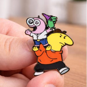 以下が含まれることがあります： 茶色のキャップとオレンジ色のパーカーを着た黄色いキャラクターが、大きな目と白いシャツ、青いショートパンツを身につけたピンクのキャラクターを肩車し、そのピンクのキャラクターが紫色のとんがり帽子をかぶった小さな緑色の生き物を持っている金属製のエナメルピン。
