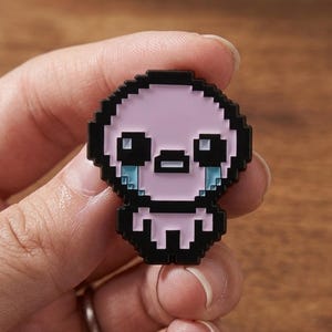 May include: A pixel art enamel pin featuring a light pink character with a round head and small body. The character has large black eyes, a small white mouth, and blue tear-shaped markings beneath the eyes, all outlined in black metal.