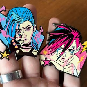 May include: Two enamel pins featuring stylised anime-inspired characters. The left pin depicts a character with blue hair and the text "Pow" in pink. The right pin shows a character with pink and blue hair, facial bandages, and yellow lightning bolts.