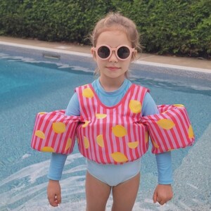 May include: A swim vest with arm floats featuring a pink and red vertical striped pattern with yellow lemon graphics. The vest is worn over a light blue long-sleeve swimsuit, paired with round, light pink-framed sunglasses with dark lenses.