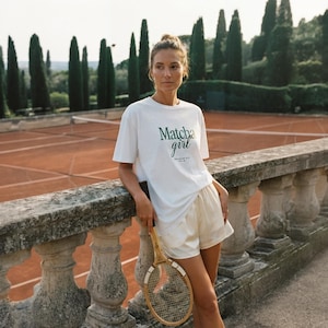 Puede incluir: Una camiseta blanca de manga corta con el texto verde oscuro "Matcha girl" estampado en el pecho, combinada con pantalones cortos de satén color crema. Una raqueta de tenis de madera con empuñadura blanca se sostiene en una mano, apoyada en una balaustrada de piedra.