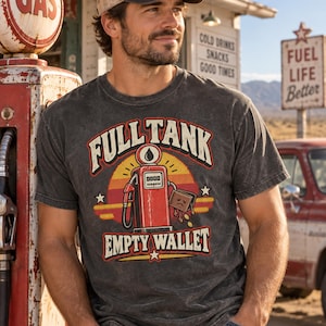 Könnte beinhalten: Ein anthrazitfarbenes Kurzarm-T-Shirt mit einer Grafik einer roten Vintage-Zapfsäule, einem traurigen Geldbeutel und dem Text "FULL TANK" sowie "EMPTY WALLET" in Retro-Schrift vor einem stilisierten orange-gelben Sonnenuntergang.