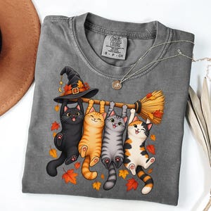 May include: A charcoal gray t-shirt featuring a graphic of four cats hanging from a broomstick. The cats are black, orange tabby, gray tabby, and calico. A black witch hat sits on the broom handle, and orange autumn leaves surround the cats.