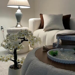 May include: A white artificial cherry blossom tree in a ribbed ceramic pot, featuring dark branches and small white flowers with yellow centers. The tree is positioned next to a round white textured ottoman, a white sofa with brown pillows, and a coffee table.