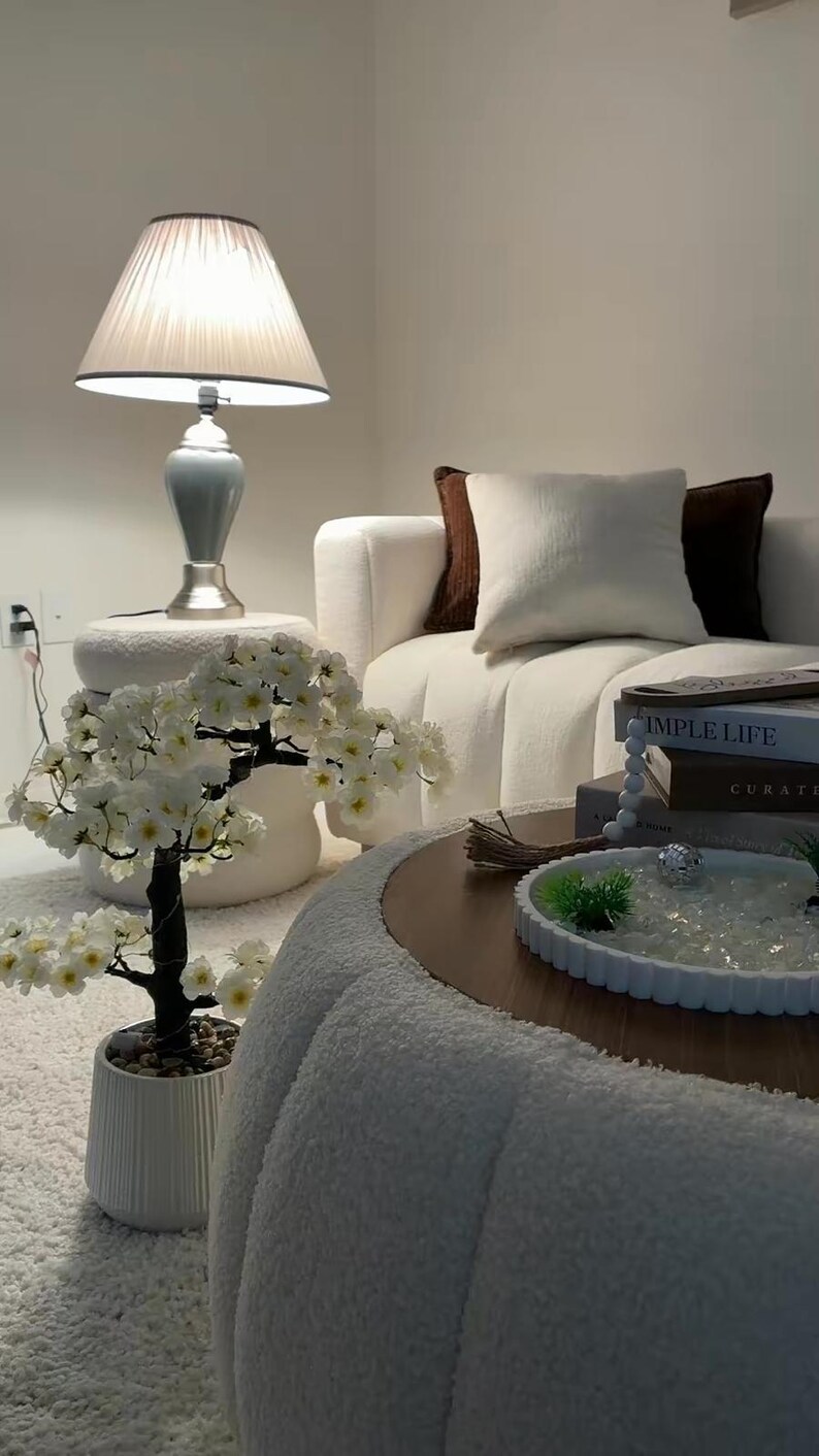 May include: A white artificial cherry blossom tree in a ribbed ceramic pot, featuring dark branches and small white flowers with yellow centers. The tree is positioned next to a round white textured ottoman, a white sofa with brown pillows, and a coffee table.