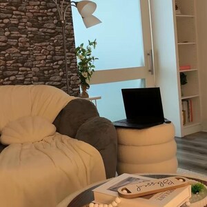 May include: A round, cream-colored, textured ottoman serves as a coffee table, topped with a stack of books, a small decorative wooden tray with the word Blessed, and a string of wooden beads. A cream-colored sofa and a black laptop are nearby.