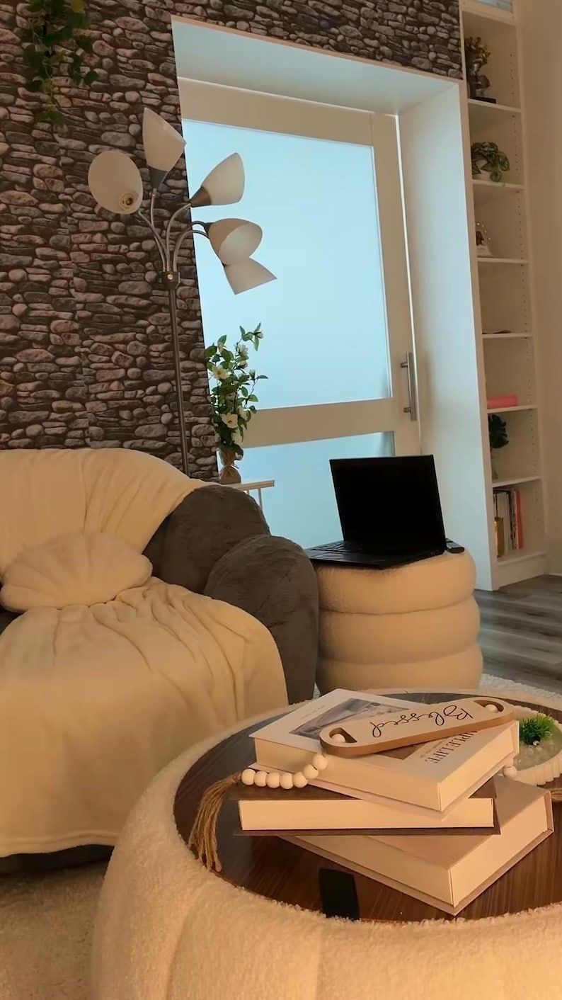 May include: A round, cream-colored, textured ottoman serves as a coffee table, topped with a stack of books, a small decorative wooden tray with the word Blessed, and a string of wooden beads. A cream-colored sofa and a black laptop are nearby.