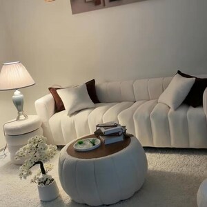 May include: A white channel-tufted sofa with dark brown and white throw pillows, a round white ottoman coffee table with a wooden top, a white side table with a lamp, a small artificial white flower tree, and wall art with geometric shapes.