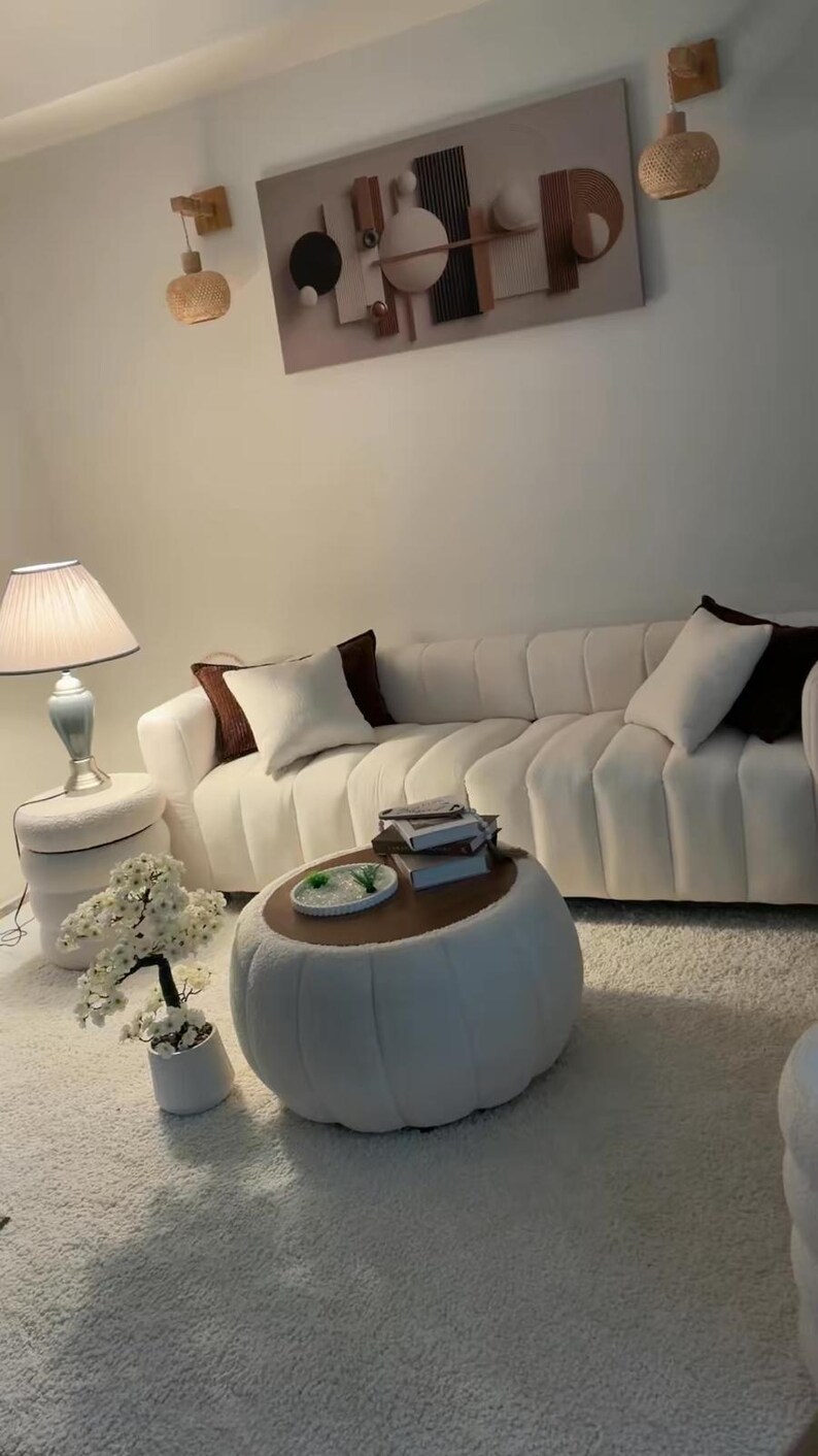 May include: A white channel-tufted sofa with dark brown and white throw pillows, a round white ottoman coffee table with a wooden top, a white side table with a lamp, a small artificial white flower tree, and wall art with geometric shapes.