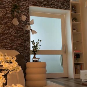 May include: A white artificial cherry blossom tree in a ribbed pot, a cream-colored cylindrical ottoman with horizontal tufting, a floor lamp with four white conical shades, and a tall white shelving unit against a stone-patterned wall.