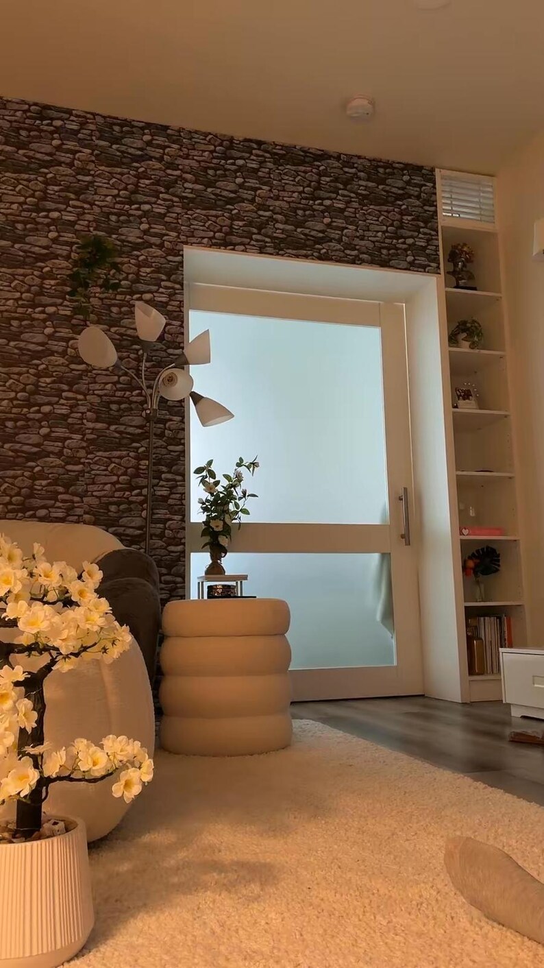 May include: A white artificial cherry blossom tree in a ribbed pot, a cream-colored cylindrical ottoman with horizontal tufting, a floor lamp with four white conical shades, and a tall white shelving unit against a stone-patterned wall.