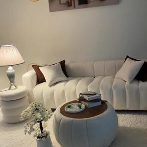 May include: A white channel-tufted sofa with dark brown and white throw pillows, a round white textured ottoman coffee table with a wooden top, a matching white side table, a table lamp, a potted white floral tree, and wall art.