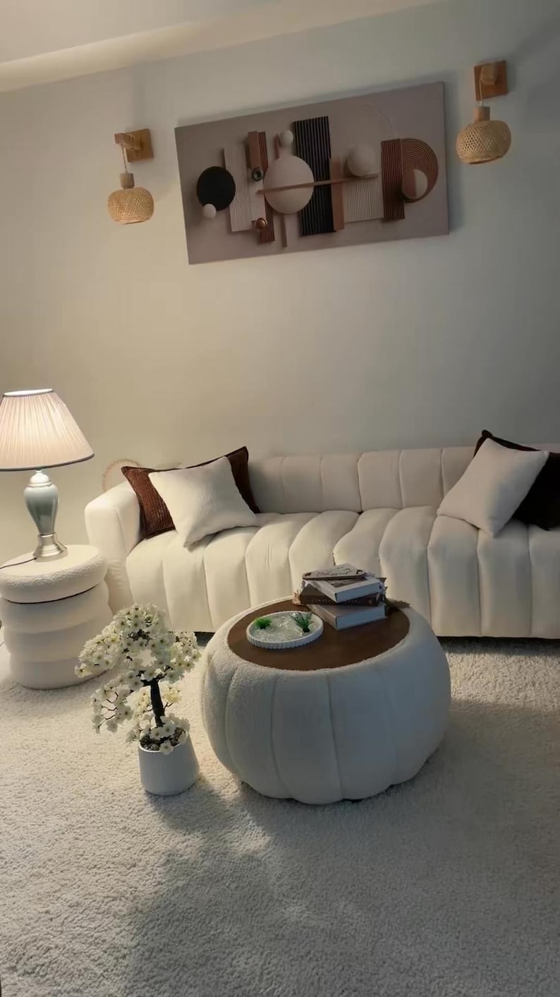 May include: A white channel-tufted sofa with dark brown and white throw pillows, a round white textured ottoman coffee table with a wooden top, a matching white side table, a table lamp, a potted white floral tree, and wall art.