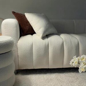 May include: A white textured sofa with vertical channel tufting, featuring one cream-colored square throw pillow and one dark brown ribbed throw pillow. A matching white cylindrical ottoman with a tiered design sits to the left of the sofa.