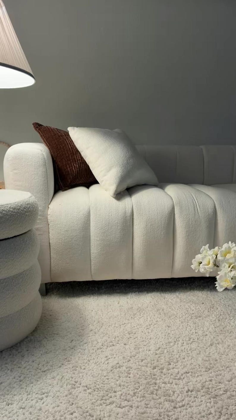 May include: A white textured sofa with vertical channel tufting, featuring one cream-colored square throw pillow and one dark brown ribbed throw pillow. A matching white cylindrical ottoman with a tiered design sits to the left of the sofa.
