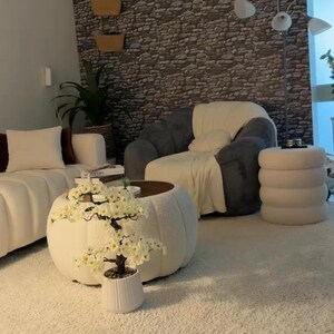 May include: A living room featuring a white tufted coffee table with a white artificial cherry blossom tree in a ribbed pot, a dark gray armchair with a white throw blanket, a white sofa, a white cylindrical stool, and a stone-patterned wall.