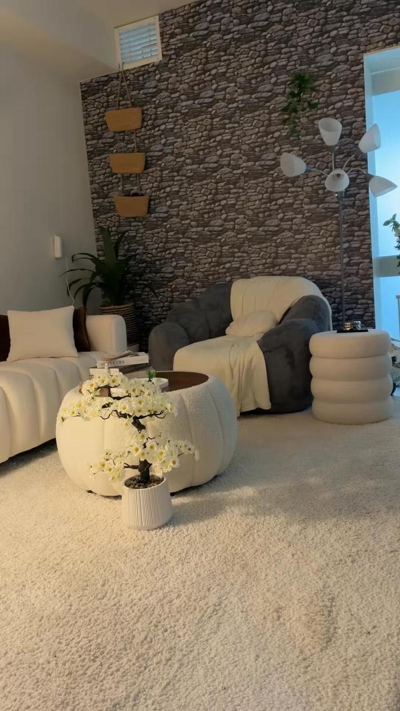 May include: A living room featuring a white tufted coffee table with a white artificial cherry blossom tree in a ribbed pot, a dark gray armchair with a white throw blanket, a white sofa, a white cylindrical stool, and a stone-patterned wall.