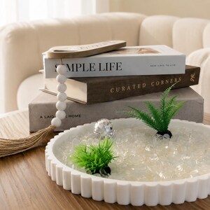 May include: A white circular tray with a fluted edge containing clear glass stones, two small green artificial plants, and a clear faceted sphere. Behind the tray are three stacked books titled Simple Life and Curated Corners with a wooden bead garland.
