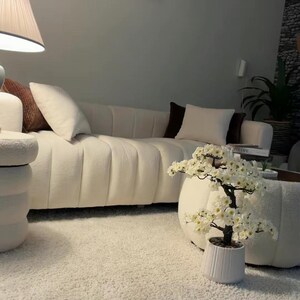 May include: A cream-colored channel-tufted sofa with white and brown throw pillows, a white textured ottoman, and a small artificial cherry blossom tree in a white fluted pot, all arranged on a thick white shag rug in a bright living space.