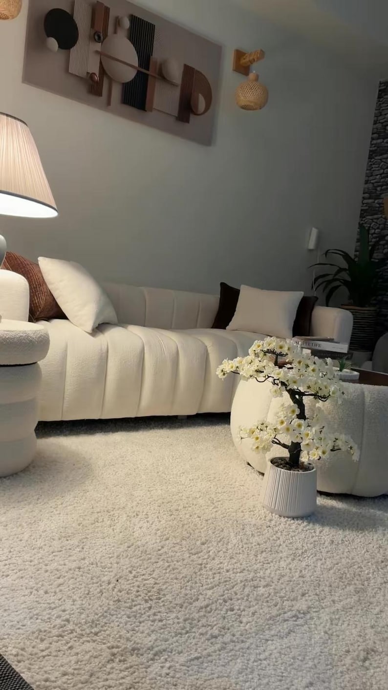 May include: A cream-colored channel-tufted sofa with white and brown throw pillows, a white textured ottoman, and a small artificial cherry blossom tree in a white fluted pot, all arranged on a thick white shag rug in a bright living space.