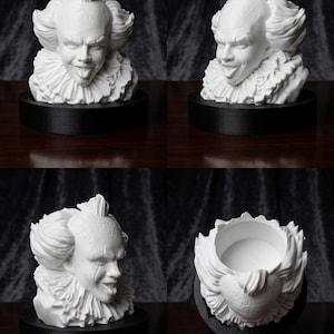 Può includere: Un busto bianco stampato in 3D di un personaggio clown con colletto a balze, caratterizzato da un'espressione facciale dettagliata con la lingua di fuori. Il busto è montato su una base nera circolare e presenta un'apertura cava in alto.