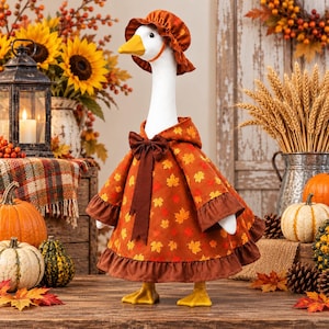 May include: A white goose figurine wearing a brown bonnet and a matching dress featuring a pattern of orange, yellow, and red autumn maple leaves. The dress has a dark brown bow at the neck and a ruffled hem. The goose has a bright yellow beak.
