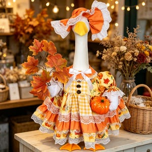 May include: A white goose figurine wearing an orange and yellow gingham dress with lace trim, a matching bonnet, and a white ribbon bow at the neck. The goose holds a cluster of orange maple leaves in one wing and a small orange pumpkin in the other.