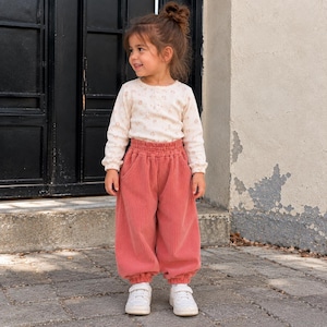 May include: A long-sleeved cream-colored shirt with a subtle floral pattern paired with loose-fitting, high-waisted, dusty rose corduroy pants featuring an elastic waistband, side pockets, and gathered cuffs, worn with white sneakers.