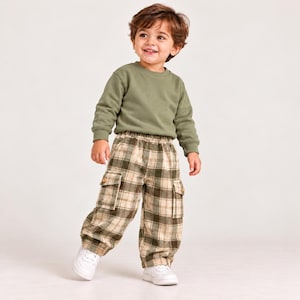 May include: A long-sleeve olive green crewneck sweatshirt paired with plaid cargo pants featuring a tan, cream, and olive green checkered pattern, an elastic waistband, two side cargo pockets with buttons, and white athletic sneakers.