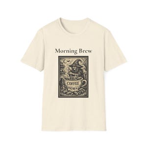 May include: A cream-coloured short-sleeve t-shirt featuring a black graphic print. The text "Morning Brew" appears above a rectangular illustration of a witch stirring a large cup labelled "COFFEE" over a fire, with a cat and moon visible.