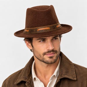 Peut inclure: Un chapeau fedora en feutre marron foncé doté d'une calotte structurée et d'un bord large. Le chapeau est orné d'une bande en cuir marron foncé texturé, entrelacée d'un cordon en cuir beige clair, créant un détail décoratif distinctif.
