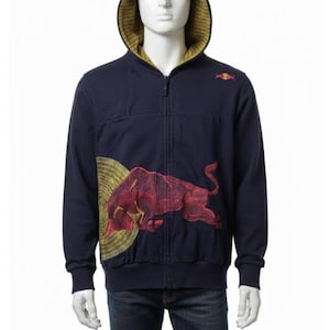 Puede incluir: Una sudadera con capucha y cremallera en azul marino oscuro que presenta un gráfico de un toro rojo en la parte delantera con un patrón circular amarillo detrás. El interior de la capucha está forrado con tela amarilla estampada. Logo pequeño.